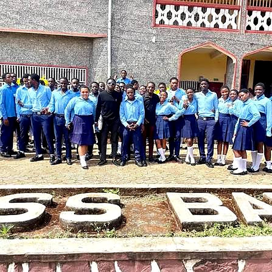 A class of PSS Bafut students in sky-blue shirts and navy skirts and trousers in front of the PSS BAFUT sign